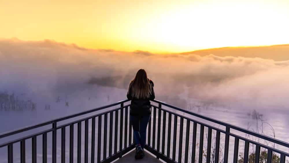Une personne se tient sur un pont surplombant une vallée enveloppée de brouillard, avec un lever de soleil en arriÚre-plan.