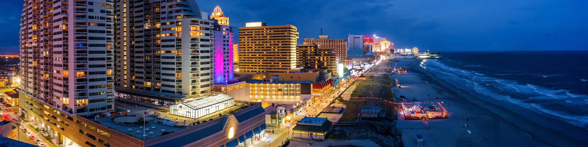 A coastal cityscape at night with illuminated buildings, a beach, and a boardwalk.