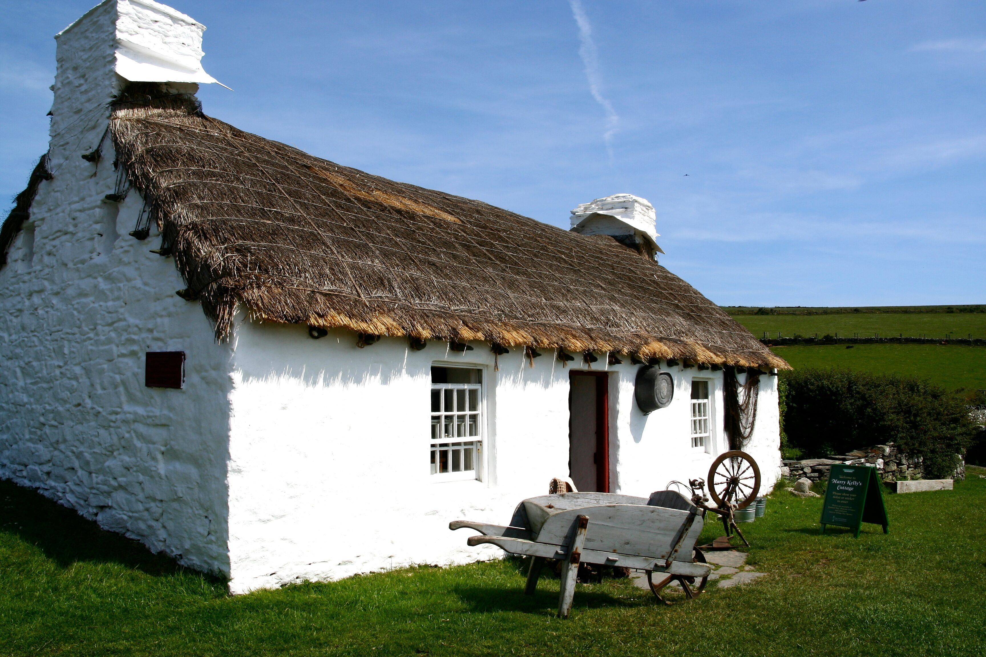 A white stone cottage with a thatched roof, a wooden wheelbarrow, and a signboard in a grassy field.