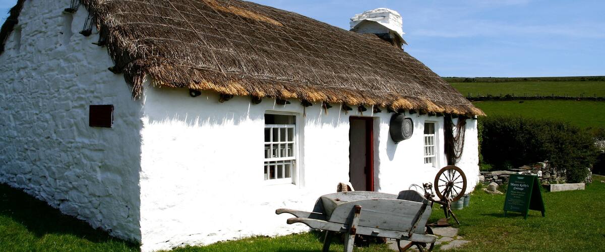 A white stone cottage with a thatched roof, a wooden wheelbarrow, and a signboard in a grassy field.