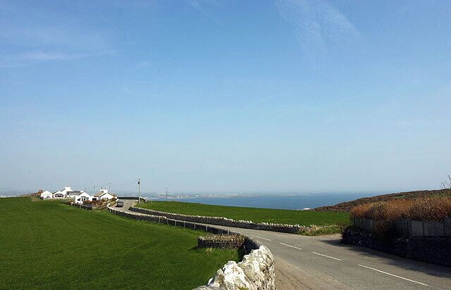 A coastal road with a stone wall, grassy fields, and white buildings in the distance.