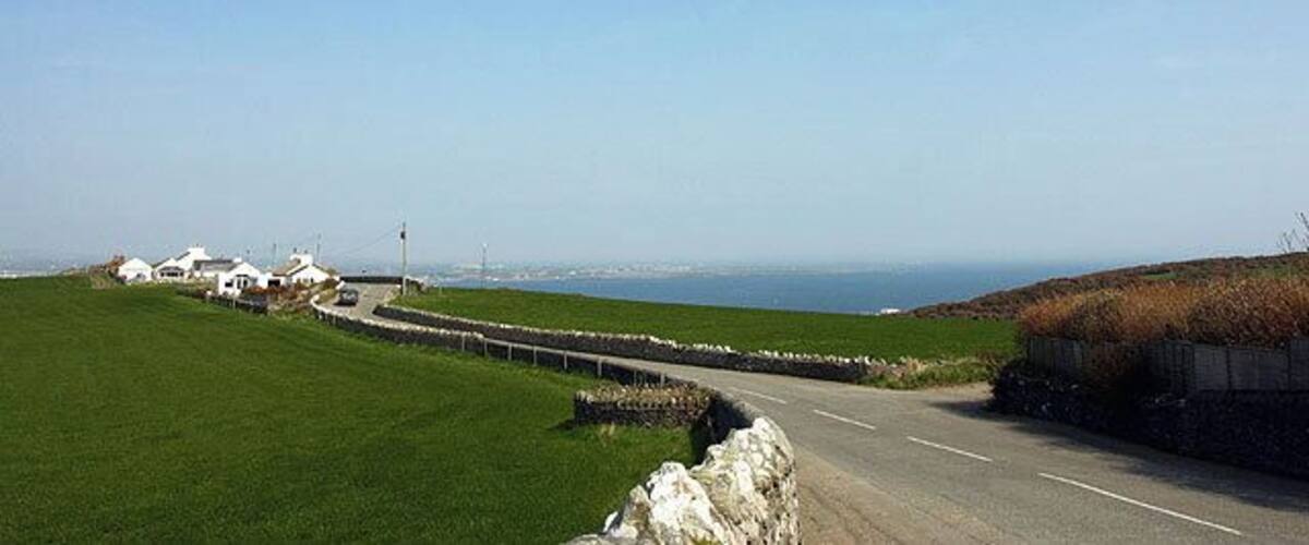 A coastal road with a stone wall, grassy fields, and white buildings in the distance.