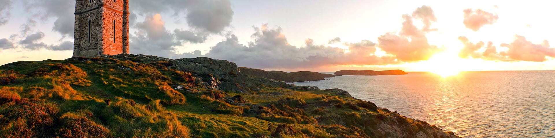 A coastal landscape with a prominent stone tower on a grassy hill, overlooking the sea at sunset.