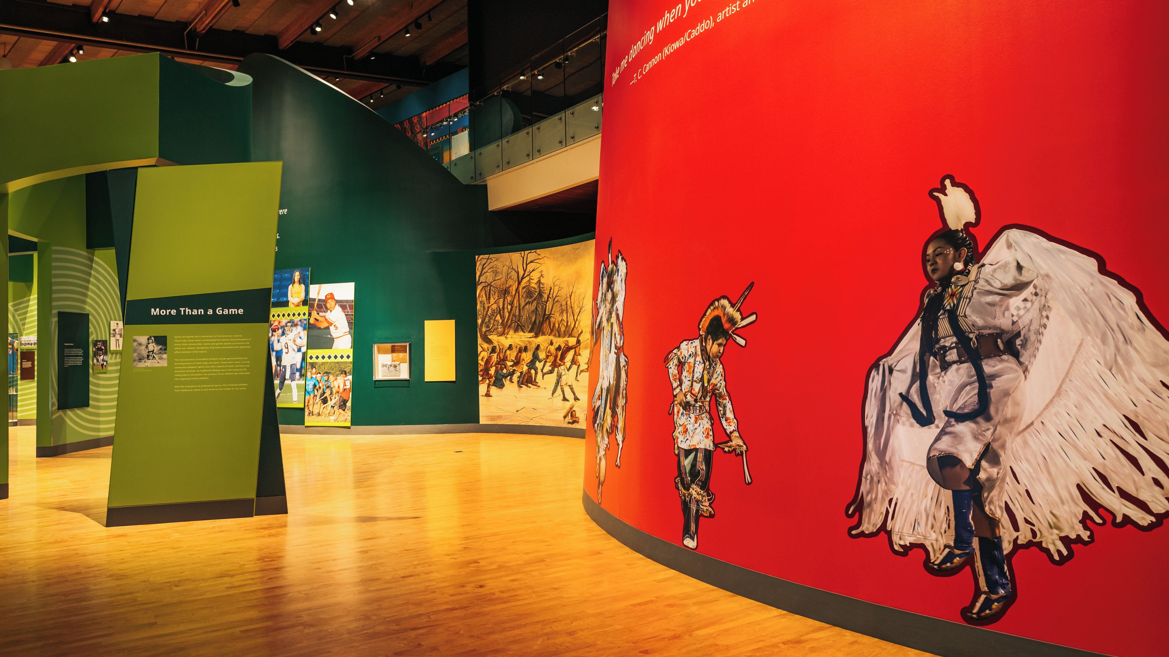 Una exhibición en un museo en interiores con paredes verdes y rojas, que presenta un gran mural de una figura nativa americana y un jugador de béisbol.