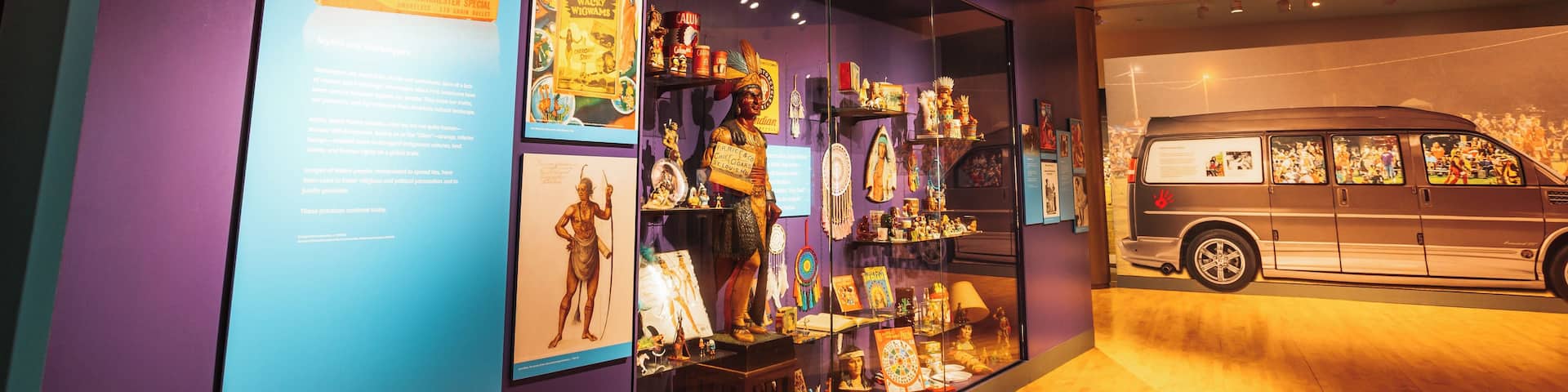 A museum exhibit with a neon sign reading 'MIS-REPRESENTATION,' displays of books and artifacts, and a van with images on its side.