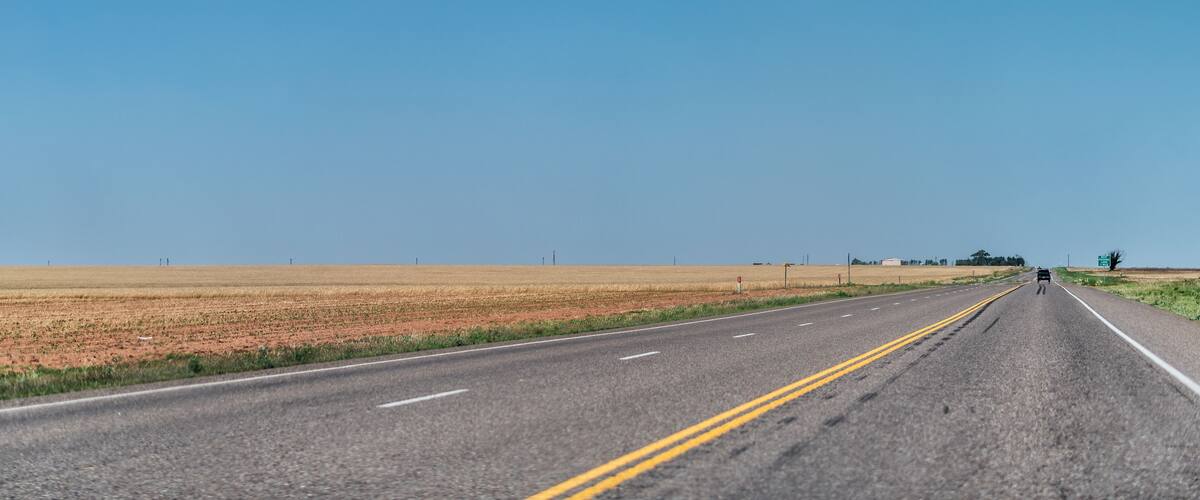 A straight road with yellow dividing lines, flanked by open fields under a clear blue sky.