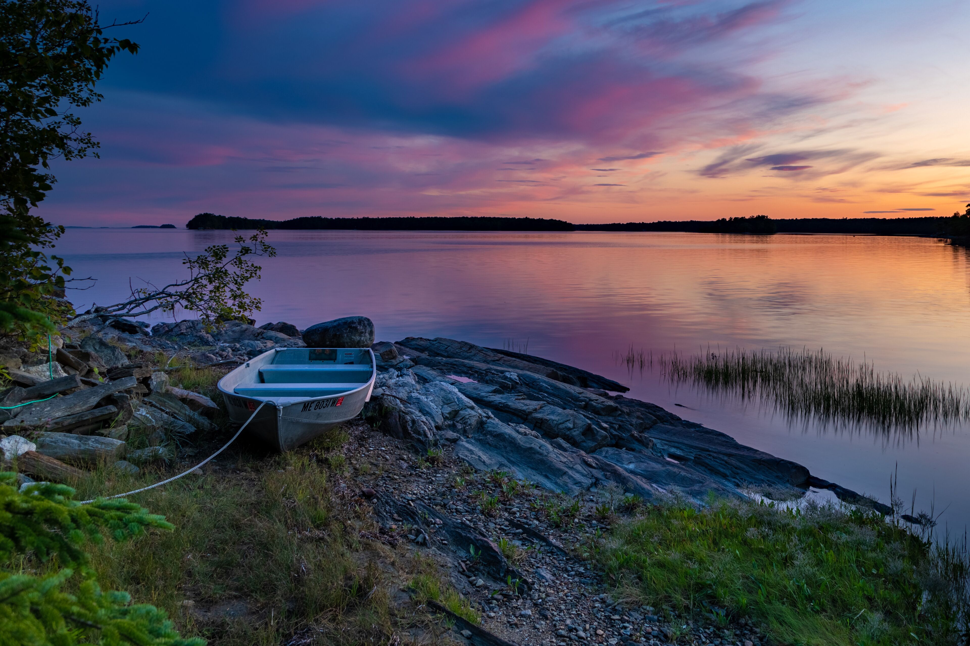 A small boat on a rocky shore with a calm lake and a colorful sunset in the background.