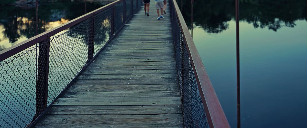 A suspension bridge over a calm body of water with a forested landscape in the background.