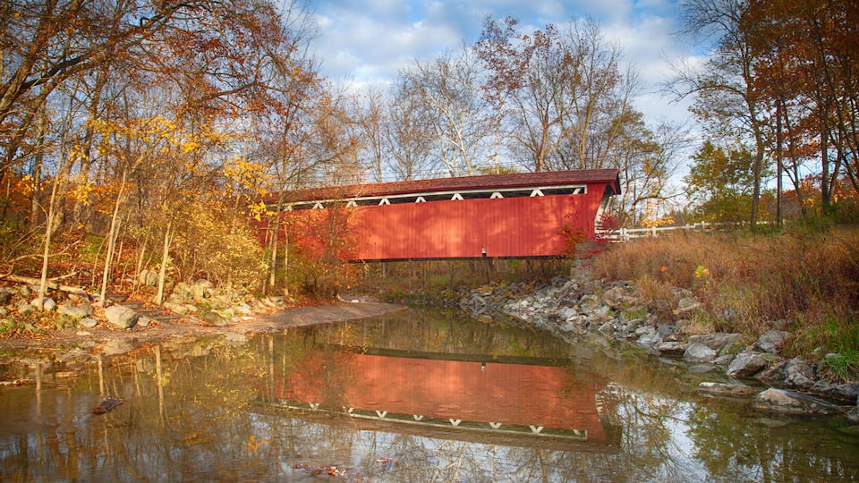 A red covered bridge over a calm river with autumn foliage in the background.