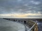 A coastal pier with a railing, waves crashing against it, and a cloudy sky.
