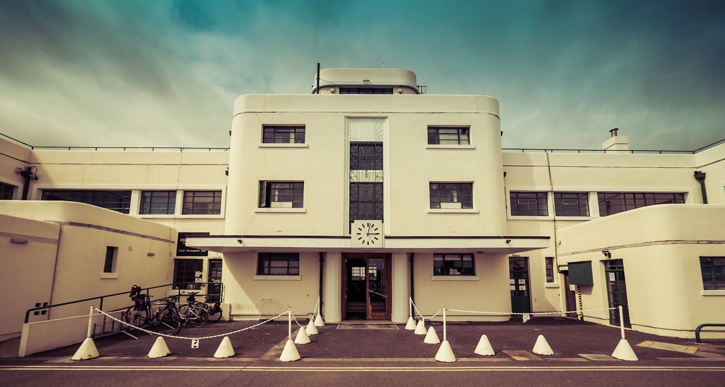 A large, white, Art Deco-style building with a central clock tower and a series of white cones marking a restricted area in front.