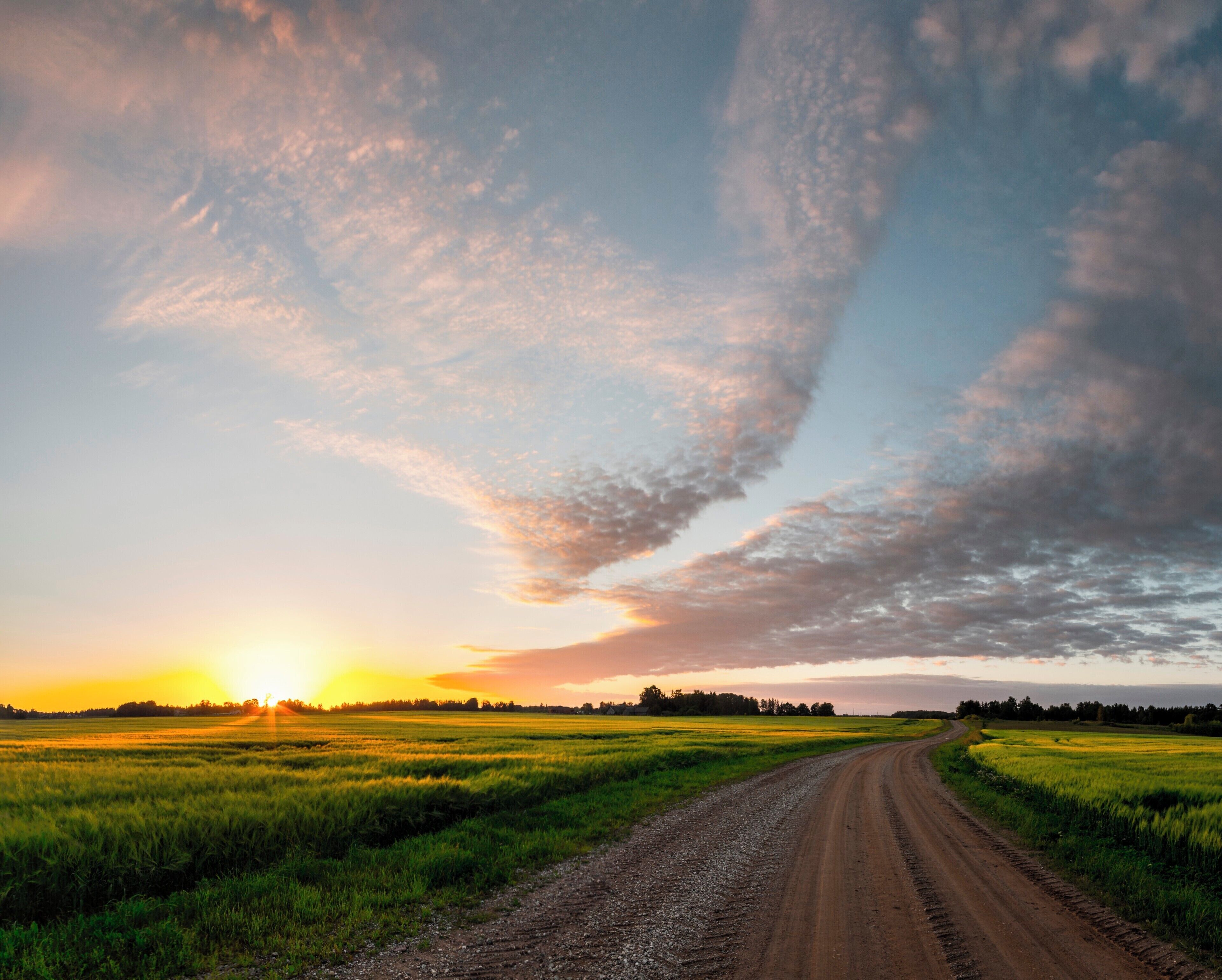 A dirt road curving through a field at sunset with a dramatic sky.