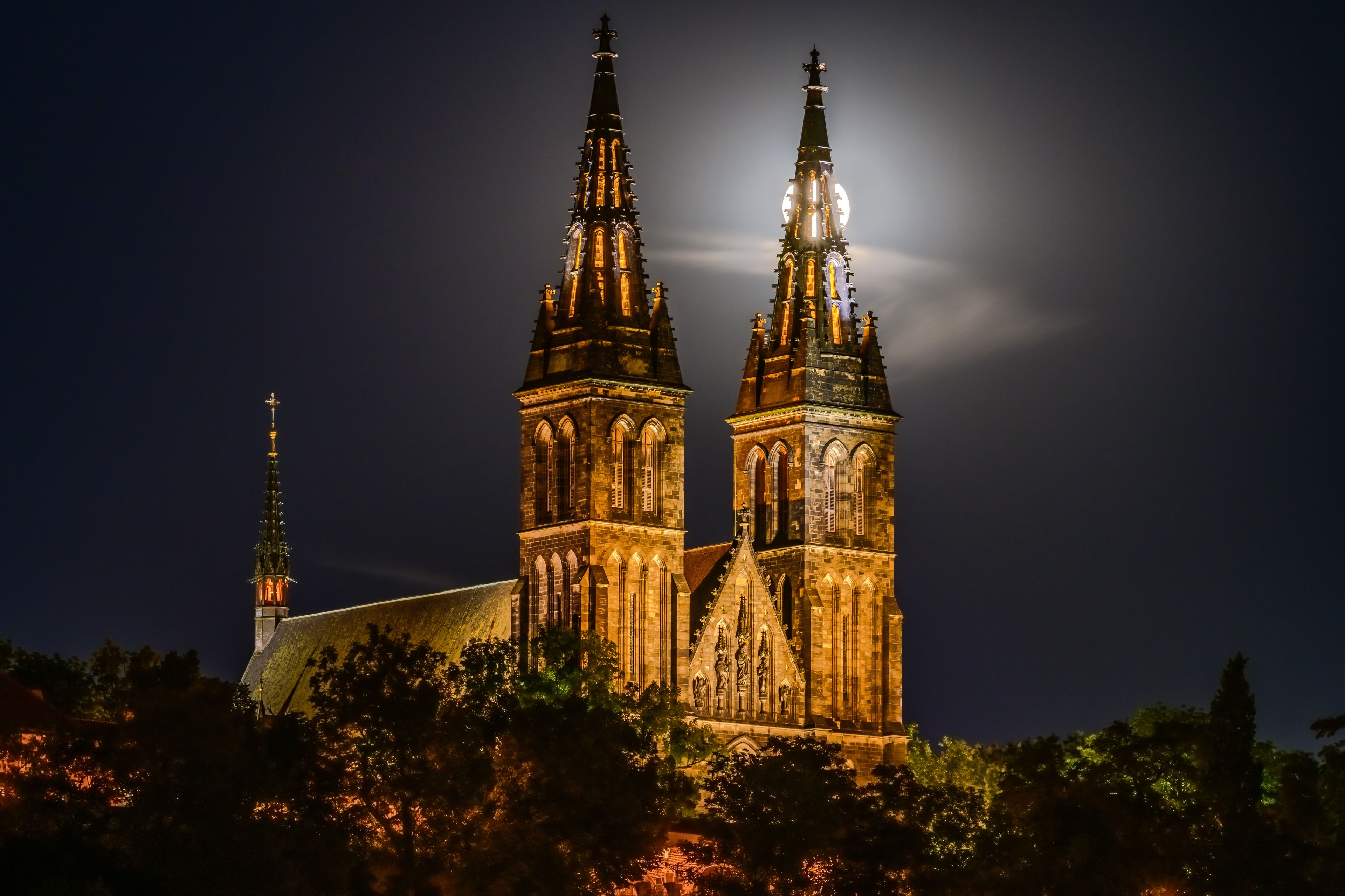 A cathedral with two tall spires, illuminated by artificial lights, under a night sky with a visible moon.