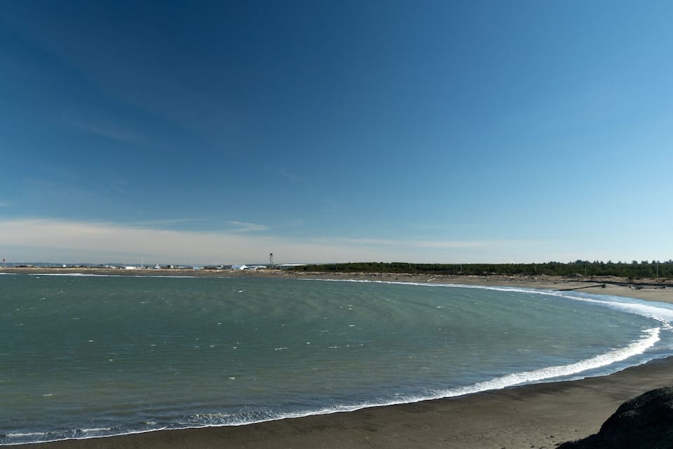 Ein Strand mit klarem, blauem Wasser, einem sandigen Ufer und einer weiter entfernt liegenden bewaldeten Gegend.