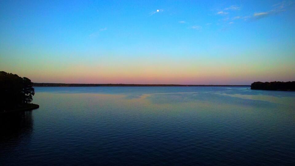A calm lake at dusk with a clear sky and a visible moon.