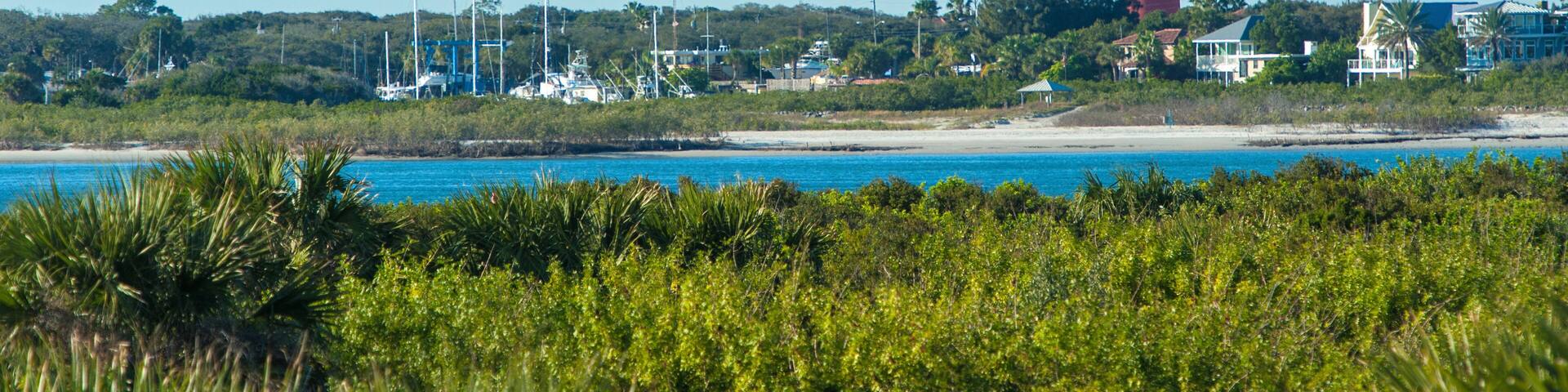 A coastal landscape with a prominent red lighthouse, clear blue sky, and lush greenery.
