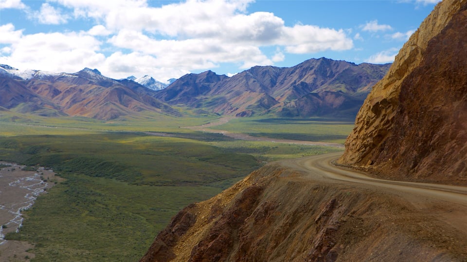 A mountainous landscape with a winding dirt road, green valleys, and snow-capped peaks.
