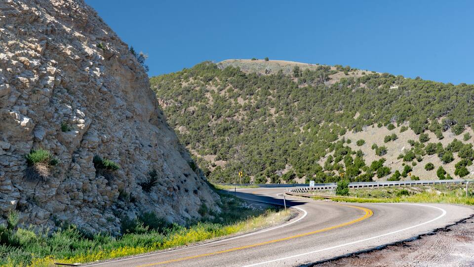 A winding mountain road with a guardrail, rocky cliffs, and sparse vegetation.