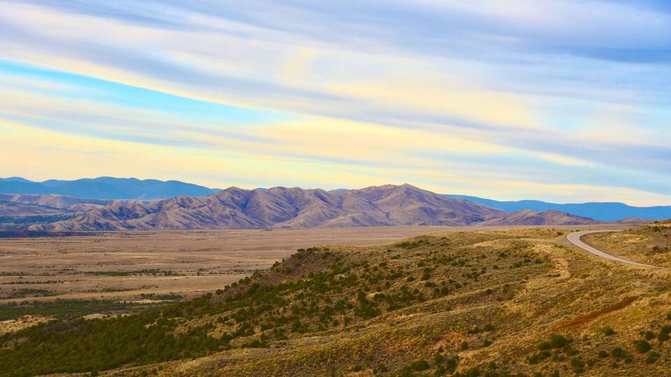A winding road through a desert landscape with distant mountains under a partly cloudy sky.