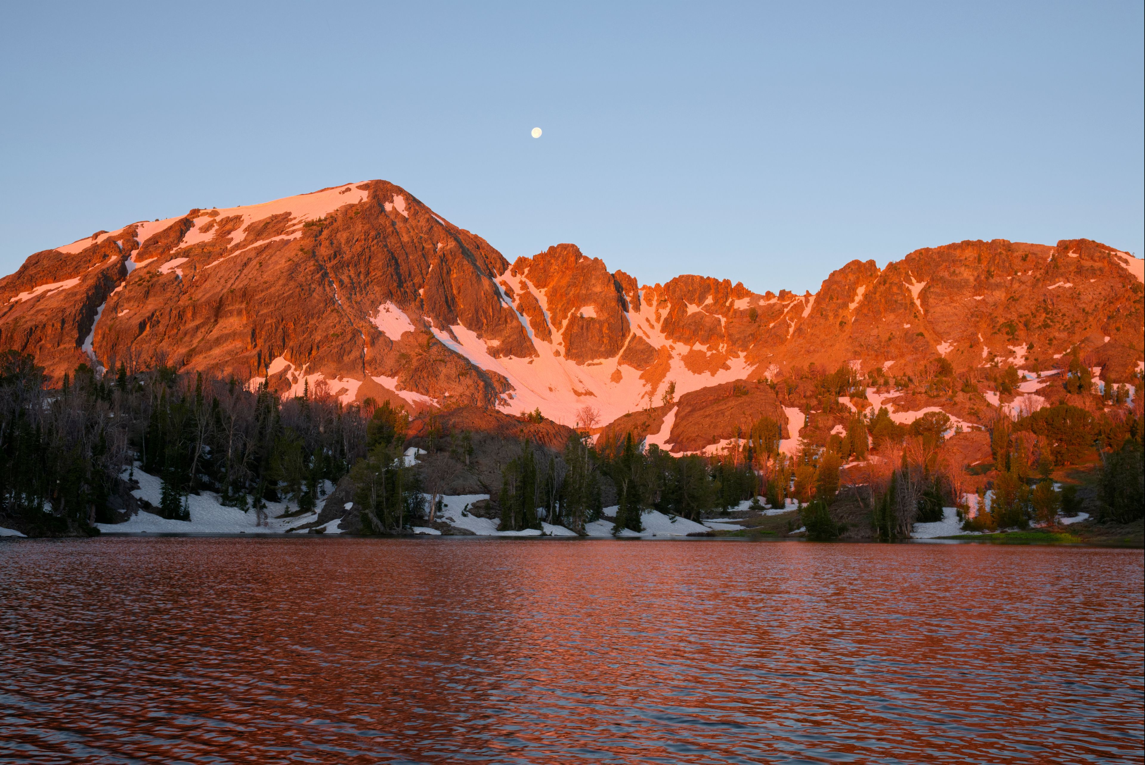 A mountain range with a lake in the foreground and a moon in the sky.