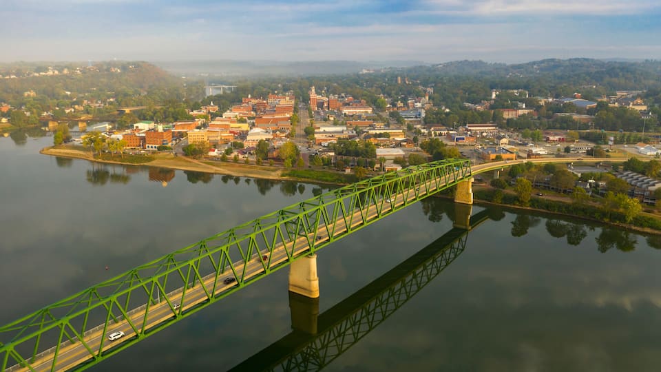 A green steel bridge spanning a calm river, surrounded by a town with various buildings and a hilly landscape in the background.