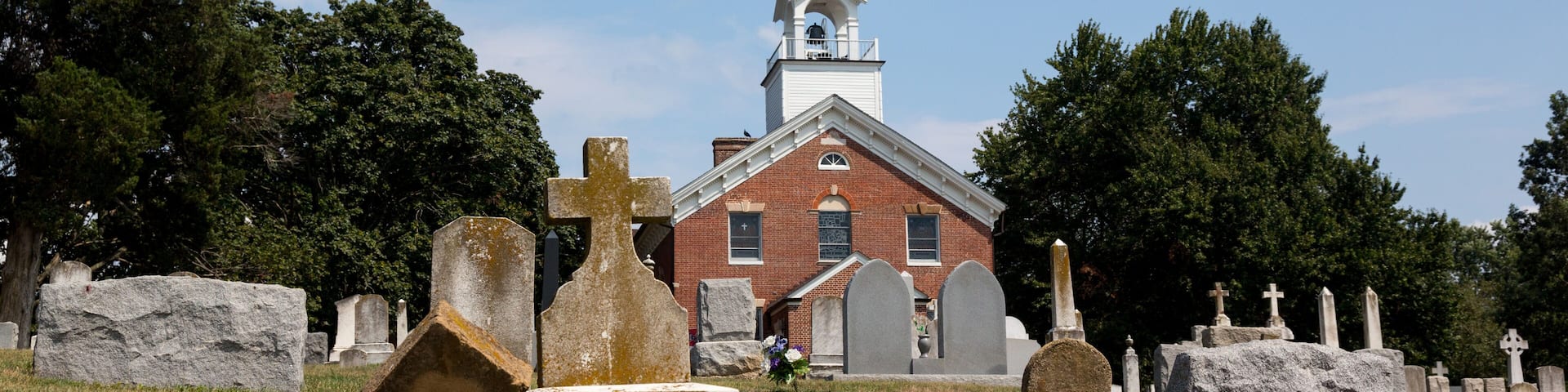 A church with a tall steeple and a brick building. There are gravestones in the foreground.