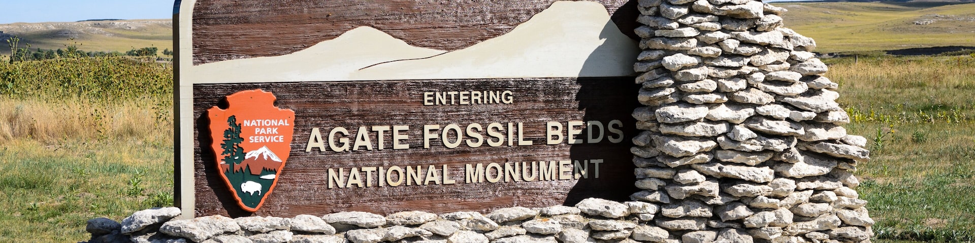 A sign for Agate Fossil Beds National Monument with a stone wall and a mountain in the background.