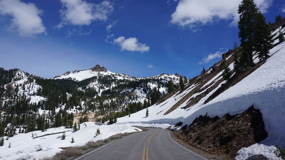 A snow-covered mountain road with a clear blue sky and a prominent peak in the distance.