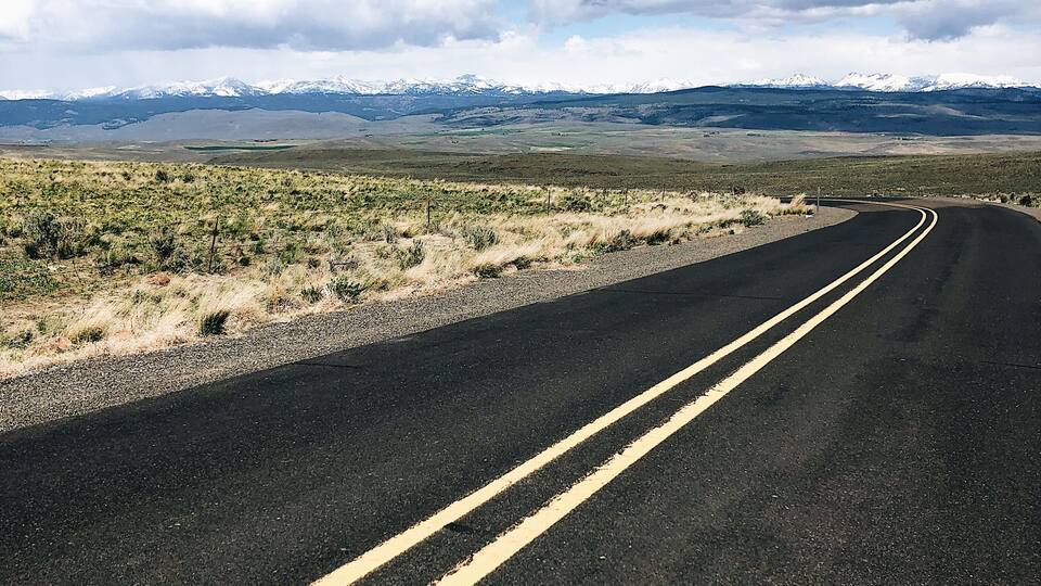 A road curving through a desert landscape with sparse vegetation and distant snow-capped mountains.