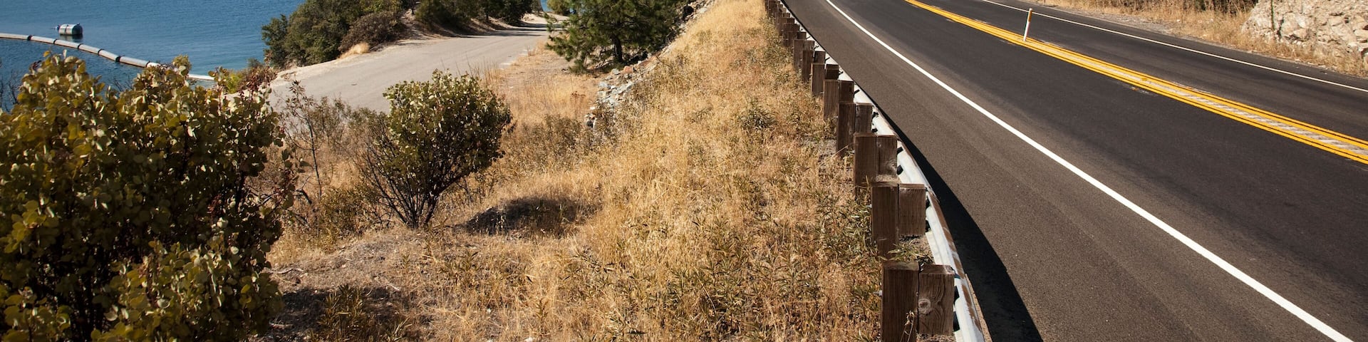 A coastal highway with a clear blue sky, a body of water, and dry hills.