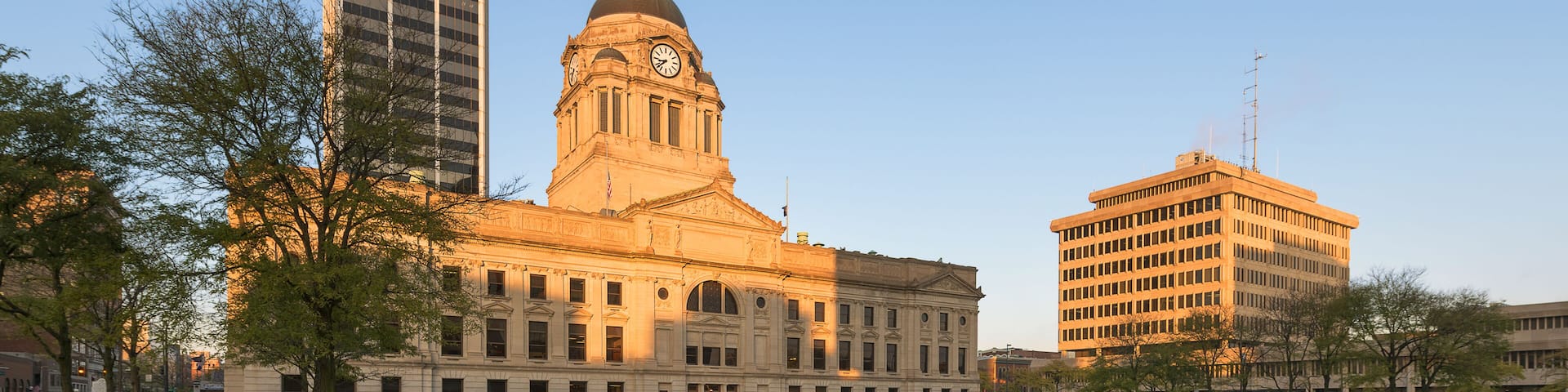 A historic building with a clock tower, surrounded by a well-maintained lawn and modern high-rise buildings.
