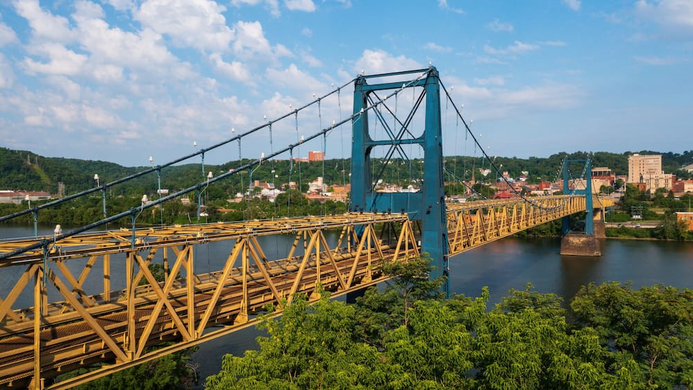 A large suspension bridge with a blue tower and yellow truss structure spanning a river, surrounded by greenery and a cityscape in the background.