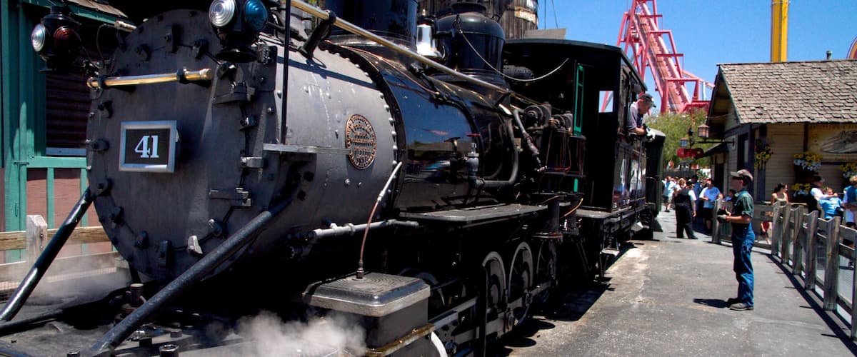 A steam locomotive numbered 41 with a dome and smokestack, parked at a station with a crowd of people and a red roller coaster in the background.