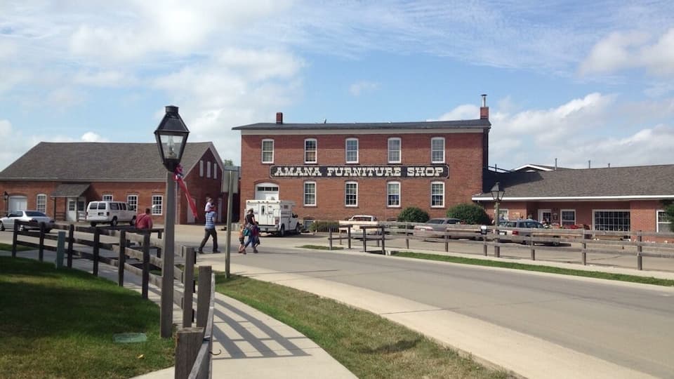 A brick building labeled "Amana Furniture Shop" with a sidewalk and street in front.