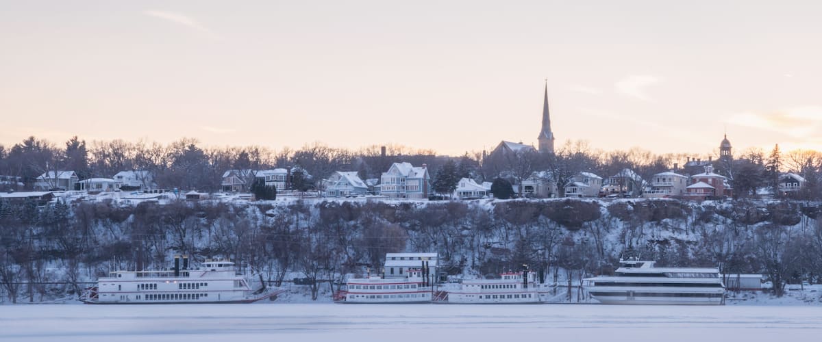 A snow-covered landscape with houses, a church steeple, and boats on a frozen river.