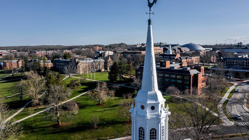 A white steeple with a rooster weather vane, surrounded by a green park and university buildings.