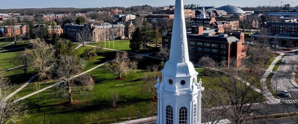 A white steeple with a rooster weather vane, surrounded by a green park and university buildings.