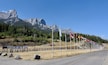 A road with a row of flags, a mountain range, and a sign indicating a ski lesson meeting place.