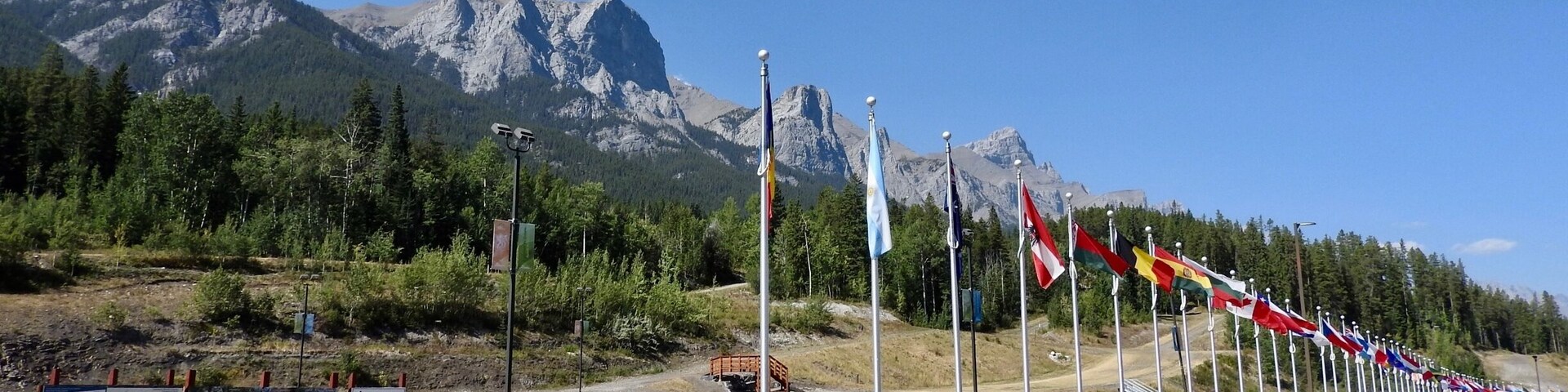 Una carretera con una hilera de banderas, una cadena montañosa y un cartel que indica el punto de encuentro para clases de esquí.