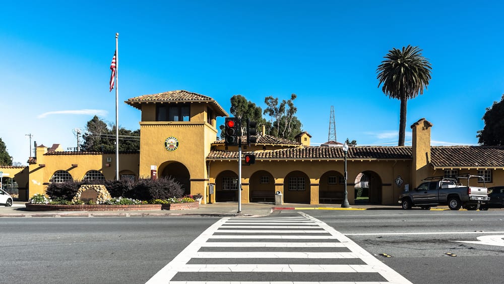 A yellow building with a clock tower, a crosswalk, and a palm tree.