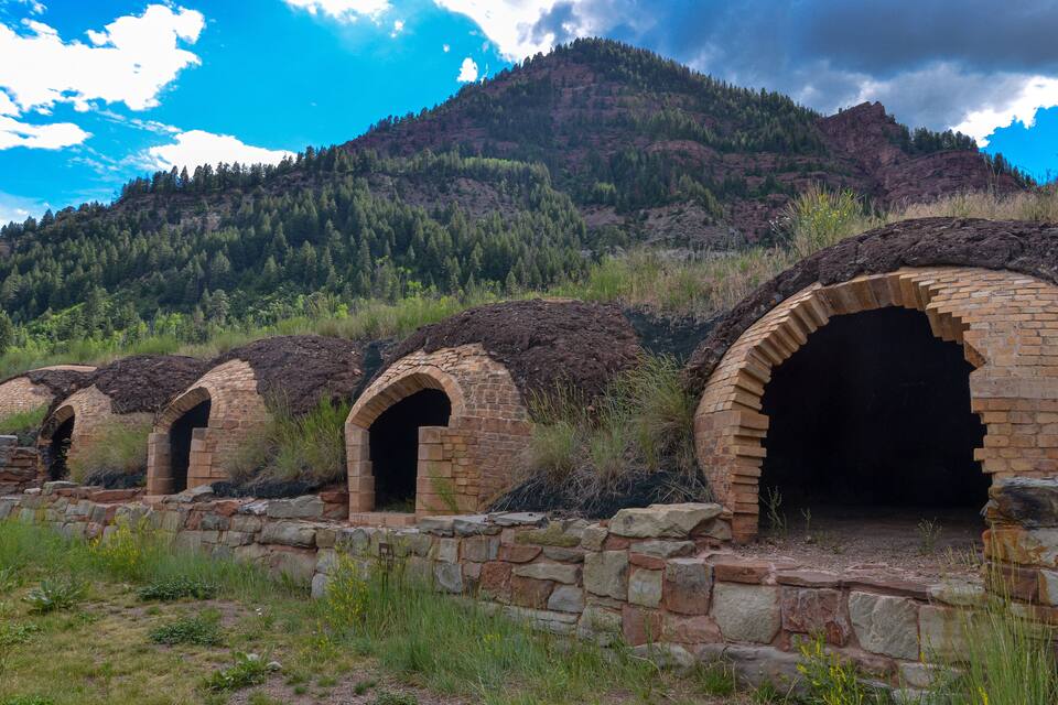 A series of brick structures with arched openings, set against a backdrop of a forested mountain under a partly cloudy sky.