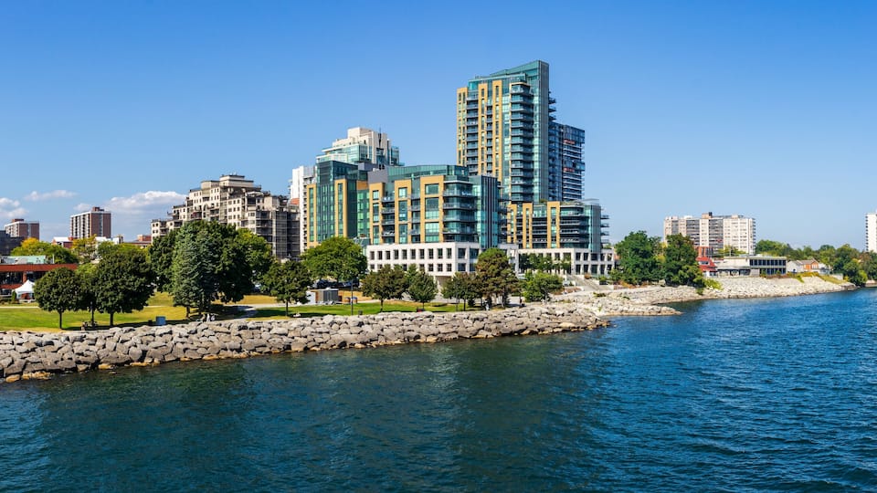 A waterfront cityscape with modern high-rise buildings, a rocky breakwater, and a clear blue sky.