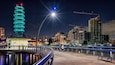 Une vue nocturne d’une ville, avec une tour illuminée, une grue de chantier et un pont où l’on voit des piétons.