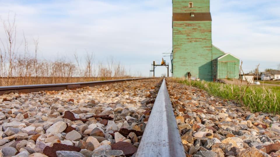 A railway track with a metal guardrail, gravel bed, and a green building with a tower in the background.