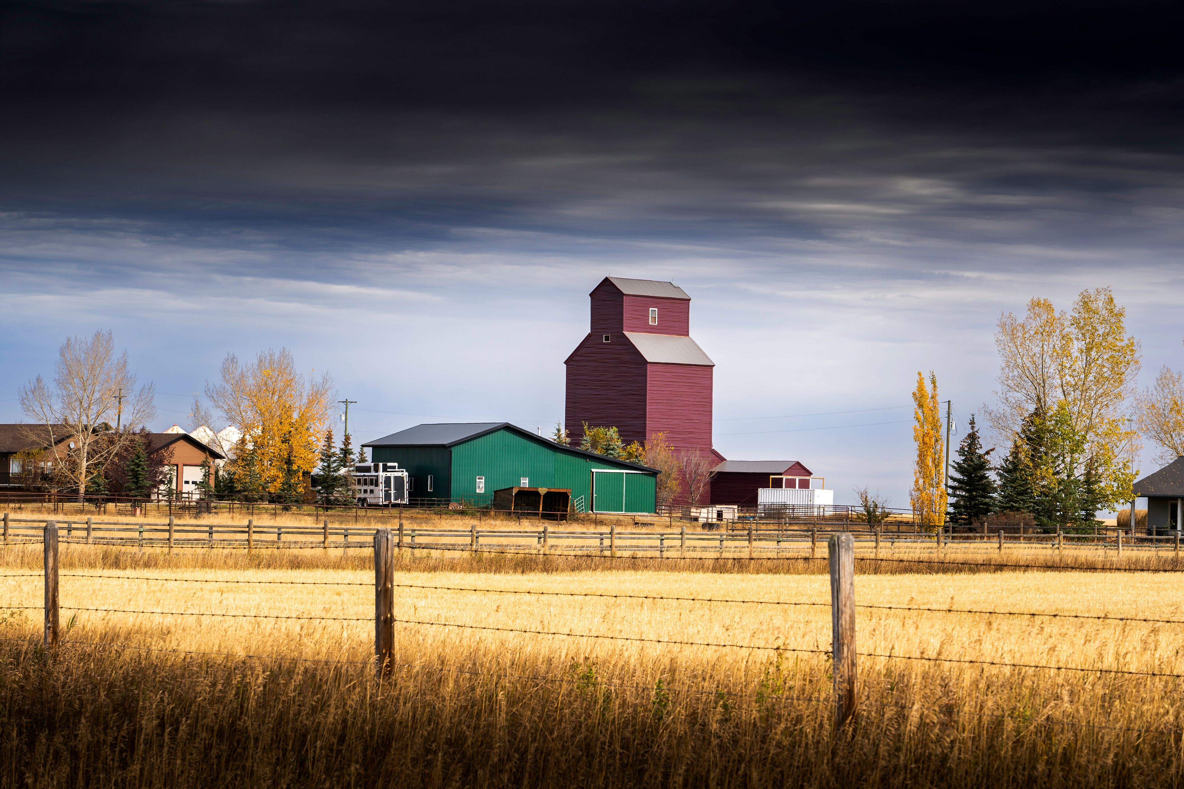 Grain elevator standing tall in a farmyard on the Alberta prairies under a dramatic sky and barbed wire fence.