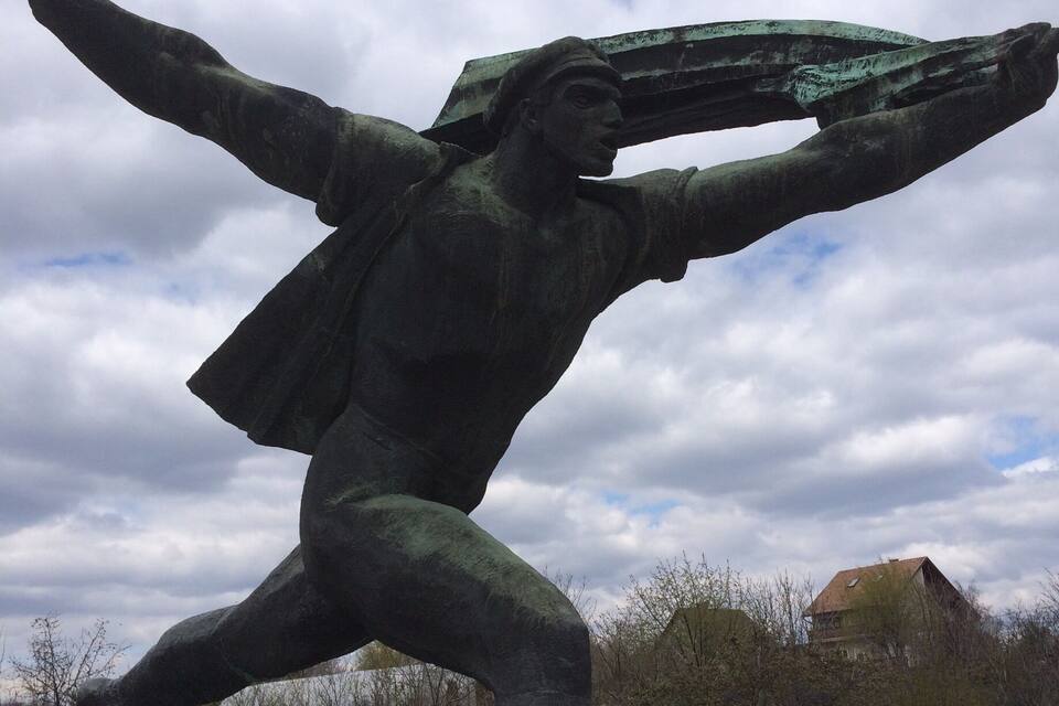 A bronze statue of a man with outstretched arms, set on a brick pedestal, with trees and a house in the background.