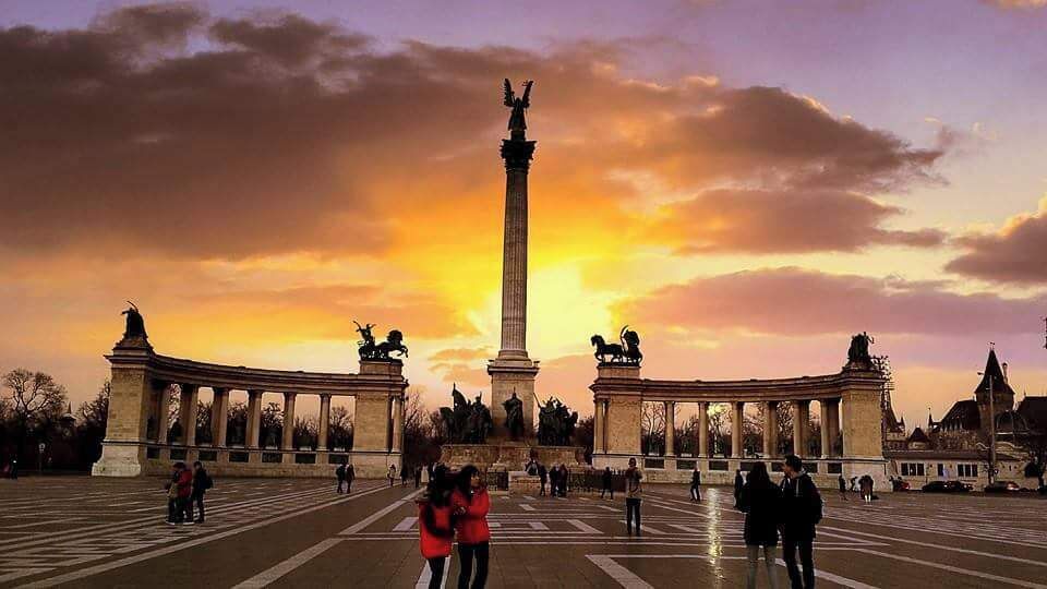 Un monumento con estatuas, rodeado por una plaza con gente y edificios al fondo.