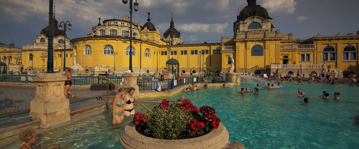 A public swimming pool with people enjoying the water, surrounded by historic yellow buildings with domed roofs.