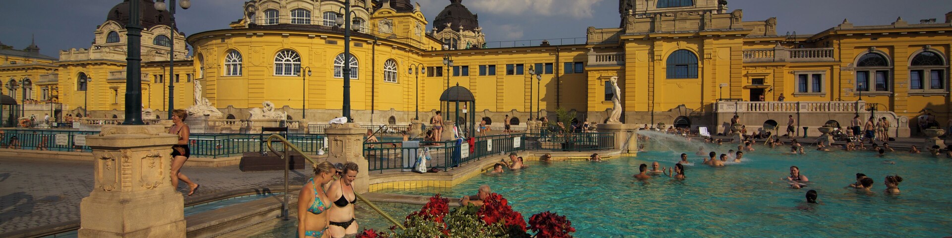 A public swimming pool with people enjoying the water, surrounded by historic yellow buildings with domed roofs.