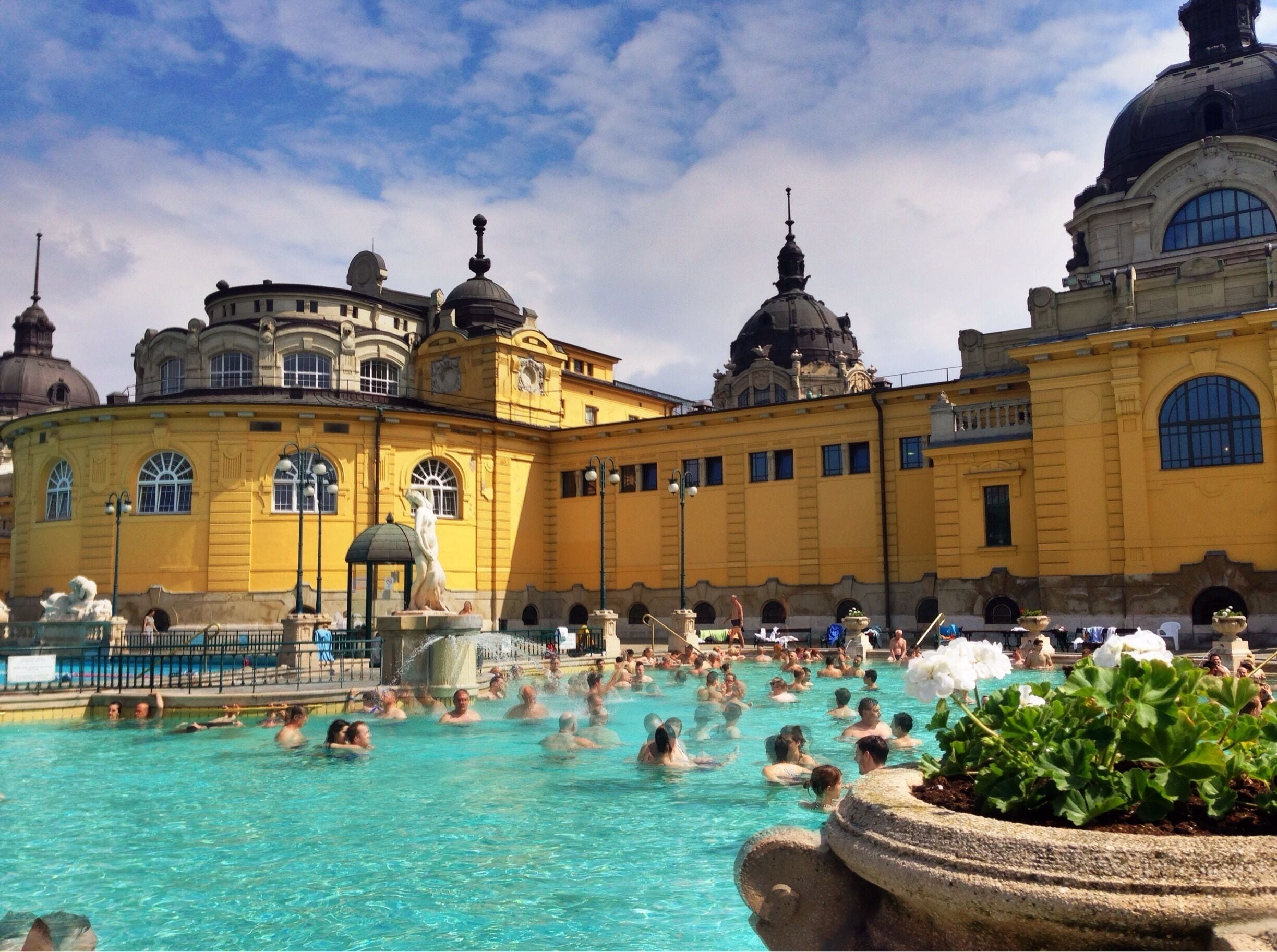 Uma grande piscina ao ar livre com pessoas aproveitando a água, rodeada por edifícios históricos amarelos com cúpulas.